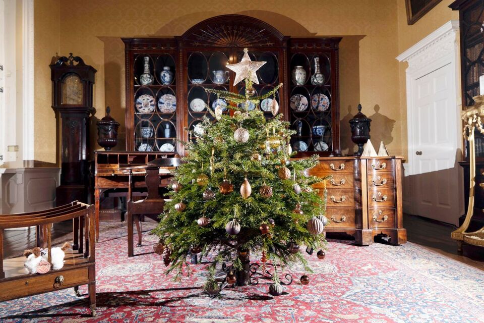 Decorated Christmas tree in an elegant room with antique wooden furniture, including a cabinet with decorative plates, a grandfather clock, and festive ornaments.